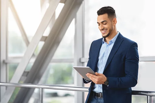 Handsome Young Arab Businessman Using Digital Tablet While Waiting At Airport Terminal, Smiling Middle Eastern Man Working Online Or Browsing Internet On Tab Computer, Enjoying Modern Technologies